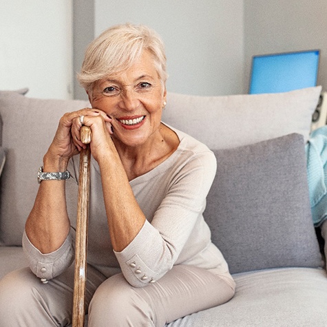 Woman wearing taupe sitting on couch leaning hands and cheek on top of walking cane